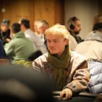 Ein Mann in einem dunkelblauen Anzug und einer weißen Krawatte sitzt an einem Pokertisch und hält zwei Spielkarten in der Hand. Er schaut konzentriert auf den Tisch, auf dem Poker