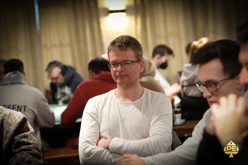 Ein Mann mit dunkler Hautfarbe und einem blauen Anzug, der Poker spielt und konzentriert auf seine Karten schaut. Er sitzt an einem grünen Pokertisch mit Chips und anderen Spielern im H