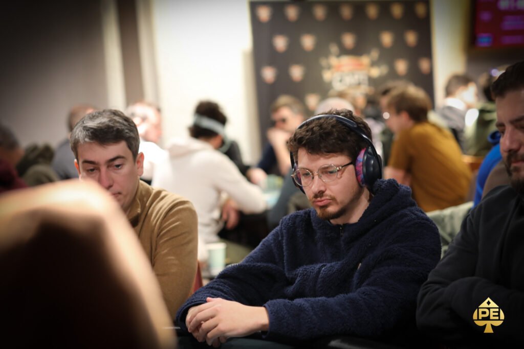 Ein Mann sitzt an einem Pokertisch, umgeben von anderen Spielern. Er hält Karten in der Hand und schaut konzentriert auf den Tisch. Die Atmosphäre ist gedämpft und spannungsg