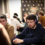 Ein Mann sitzt an einem Pokertisch, umgeben von anderen Spielern. Er hält Karten in der Hand und schaut konzentriert auf den Tisch. Die Atmosphäre ist gedämpft und spannungsg