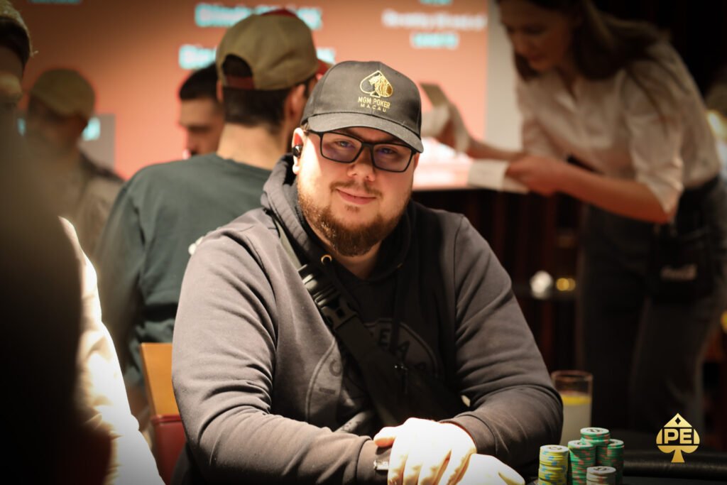 Ein Mann in einem schwarzen Anzug und einer weißen Hemdweste sitzt an einem Pokertisch und hält eine Hand mit Spielkarten. Er wirkt konzentriert und fokussiert. Im Hintergrund