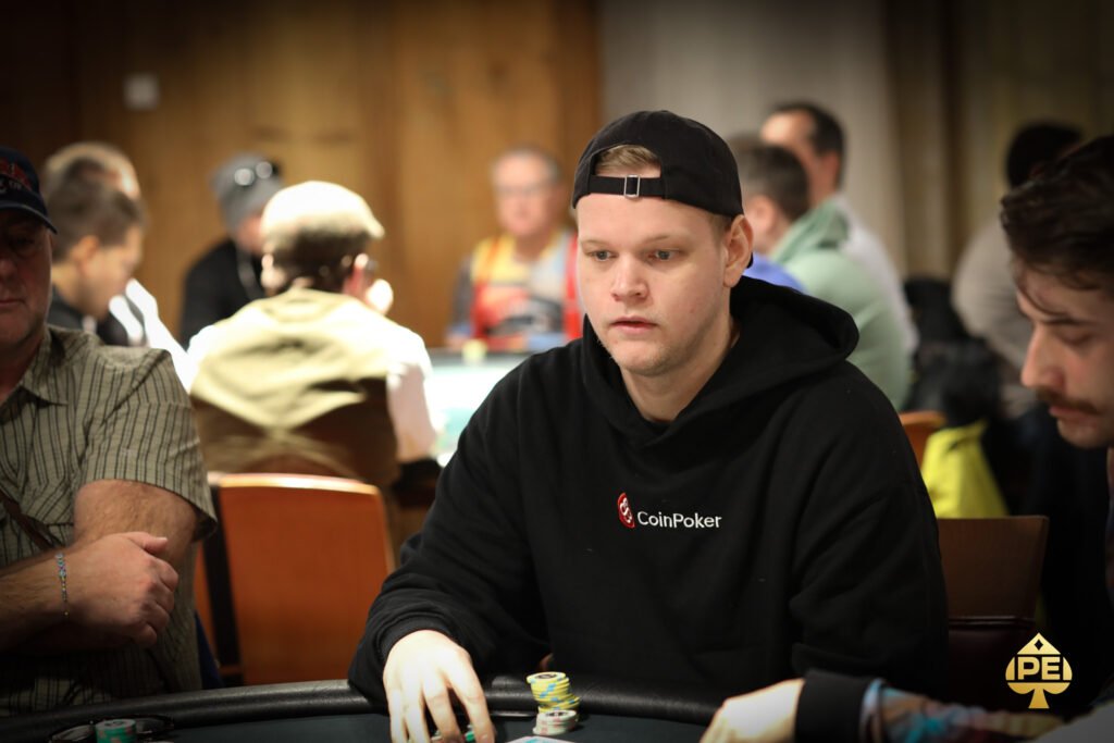 Ein professioneller Pokerspieler mit Sonnenbrille und Kappe sitzt an einem grünen Pokertisch und betrachtet konzentriert seine Karten. Im Hintergrund sind weitere Spieler und Pokerchips zu sehen