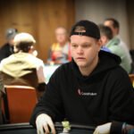 Ein professioneller Pokerspieler mit Sonnenbrille und Kappe sitzt an einem grünen Pokertisch und betrachtet konzentriert seine Karten. Im Hintergrund sind weitere Spieler und Pokerchips zu sehen
