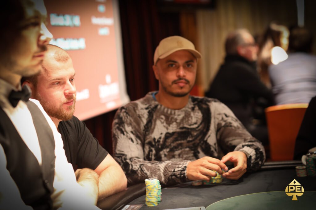 Ein Mann sitzt an einem Pokertisch in einem eleganten Casino. Er hält Karten in der Hand und schaut konzentriert auf den Tisch. Im Hintergrund sind andere Spieler und Dealer zu sehen.