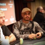 Ein Mann sitzt an einem Pokertisch in einem eleganten Casino. Er hält Karten in der Hand und schaut konzentriert auf den Tisch. Im Hintergrund sind andere Spieler und Dealer zu sehen.