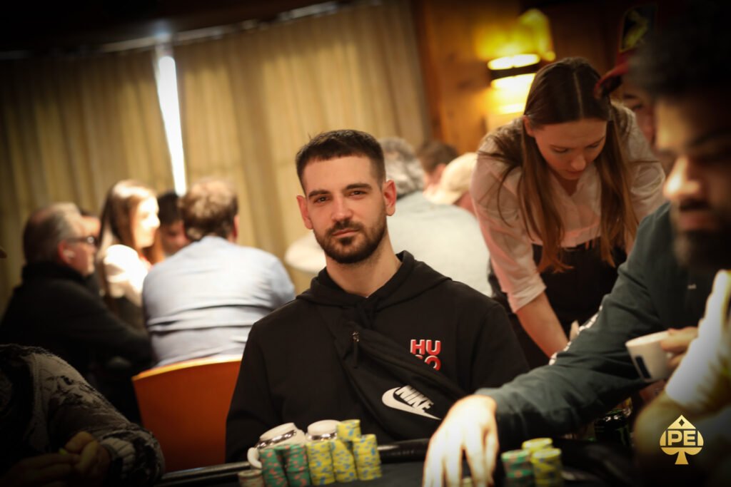 Ein Mann in einem schwarzen Anzug sitzt an einem Pokertisch mit grünem Filz. Er hält Spielkarten in der Hand und schaut konzentriert auf den Tisch. Um ihn herum sind Pokerch