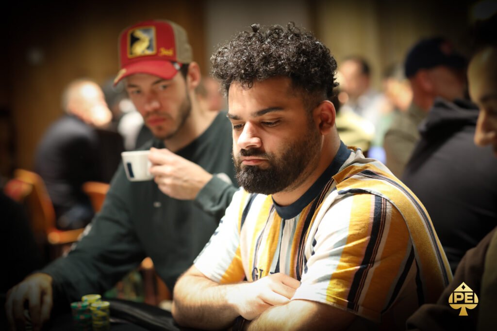 Ein Mann in einem schwarzen Anzug sitzt an einem Pokertisch, hält Karten in der Hand und schaut konzentriert auf seine Gegner. Die Spieler um ihn herum sind unscharf im Hintergrund zu