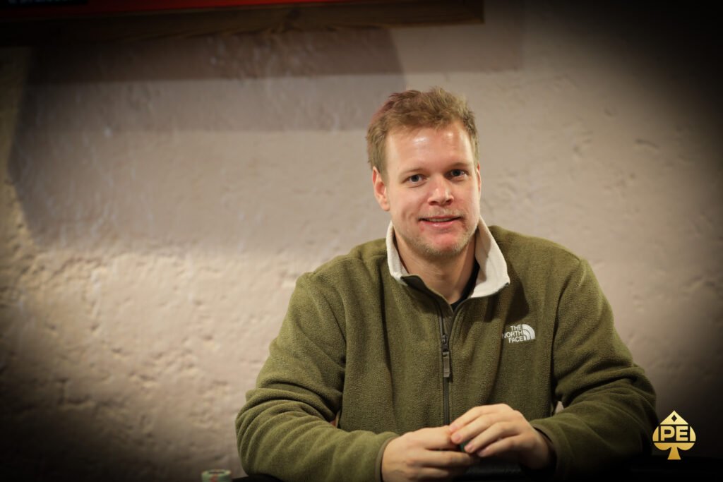 Ein Mann in einem schwarzen Anzug und einer roten Krawatte sitzt an einem Pokertisch und hält ein paar Spielkarten in der Hand, während er ernsthaft auf seine Gegner schaut.