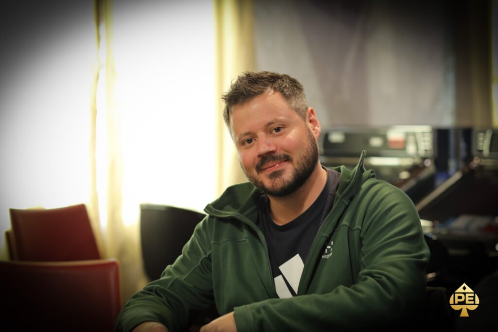 Ein Mann in einem schwarzen Anzug und einer schwarzen Sonnenbrille sitzt an einem Pokertisch und hält Karten in der Hand, während er konzentriert auf seine Gegner schaut.