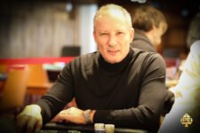 Ein Mann in schwarzer Kleidung und Sonnenbrille sitzt an einem Pokertisch und betrachtet konzentriert seine Karten. Im Hintergrund sind weitere Spieler und Chips zu sehen.
