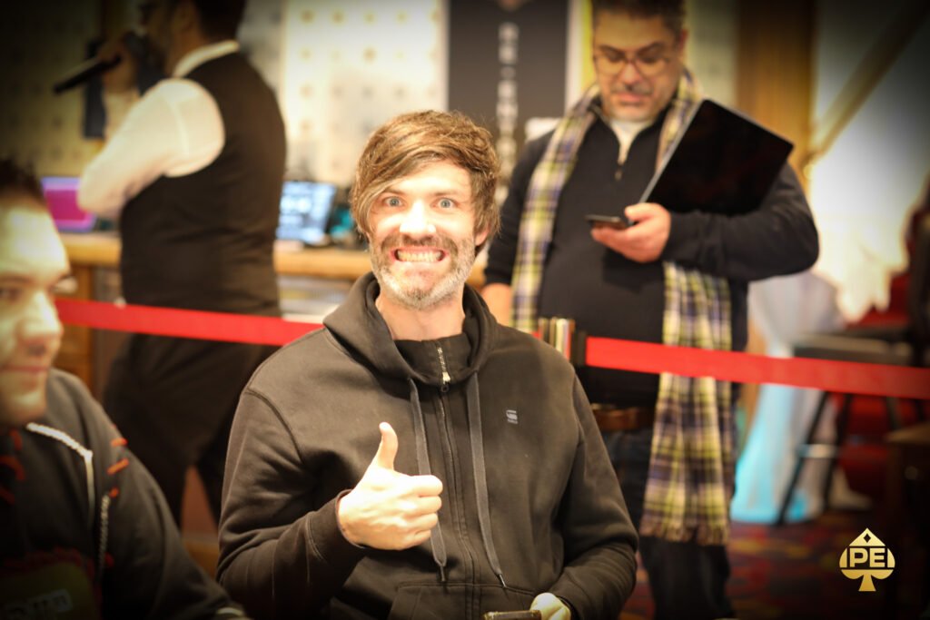 Ein Mann mit dunklen Haaren und einem schwarzen Anzug sitzt an einem Pokertisch und hält zwei Spielkarten in der Hand. Ein Stapel Pokerchips liegt vor ihm auf dem grünen Filz. Im H