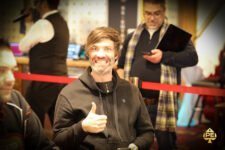 Ein Mann mit dunklen Haaren und einem schwarzen Anzug sitzt an einem Pokertisch und hält zwei Spielkarten in der Hand. Ein Stapel Pokerchips liegt vor ihm auf dem grünen Filz. Im H