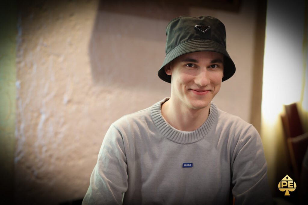 Ein Mann in einem schwarzen Anzug sitzt an einem Pokertisch und hält seine Karten in der Hand, während er konzentriert in die Kamera schaut. Im Hintergrund sind Pokerchips und Spielkarten
