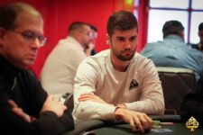 Ein älterer Mann sitzt an einem Pokertisch und hält seine Spielkarten. Er hat eine Brille auf und trägt ein Hemd mit einem Muster. Im Hintergrund sind Pokerchips und weitere Spieler zu sehen