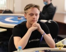 Ein Mann mit kurzen braunen Haaren und einem grauen Hemd sitzt an einem Pokertisch und hält Karten in der Hand. Er schaut konzentriert auf seine Karten und hat eine ernste Miene