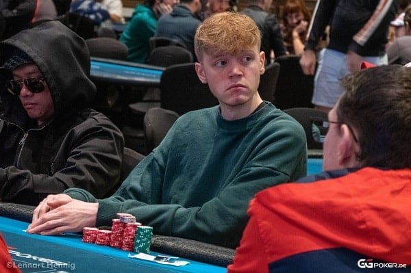 Ein Mann sitzt entspannt an einem Pokertisch und hält Karten in der Hand. Er trägt eine schwarze Brille und konzentriert sich auf das Spiel.