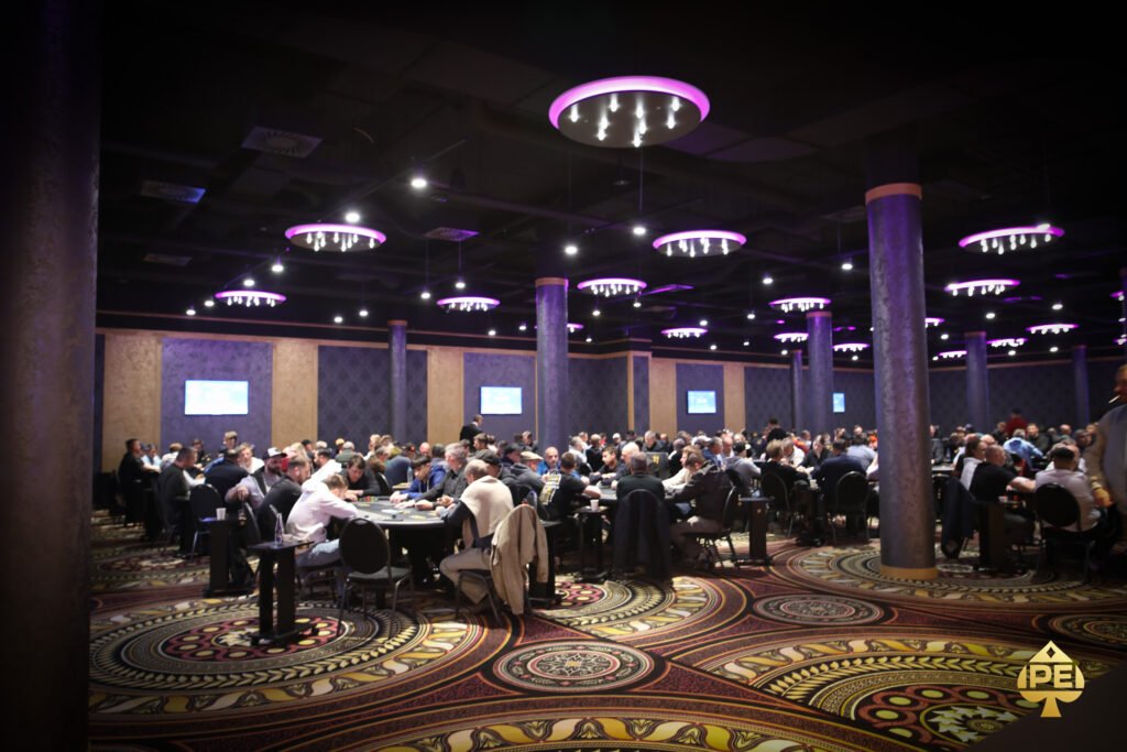 Ein Mann und eine Frau sitzen an einem Pokertisch und spielen eine Partie Poker. Der Mann hat einen Stapel Chips vor sich, während die Frau ihre Karten betrachtet. Im Hintergrund sind weitere Spieler und eine elegante