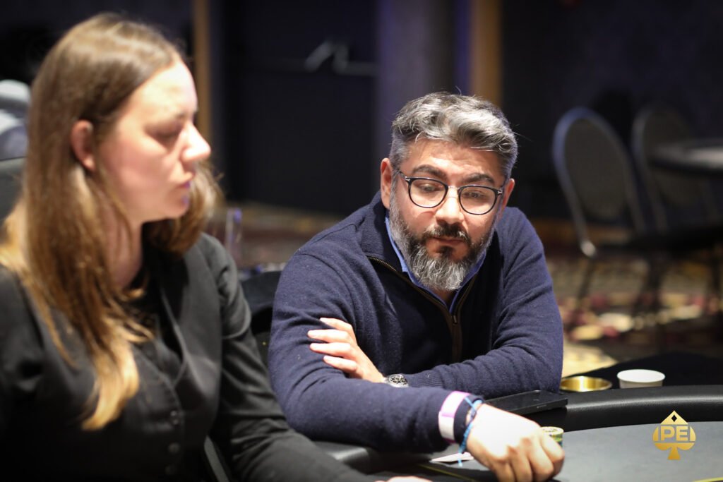 Ein Mann in einem Anzug und mit einer Sonnenbrille spielt Poker an einem Tisch mit grünem Filz. Er hält Karten in der Hand und schaut konzentriert auf den Tisch. Andere Spieler