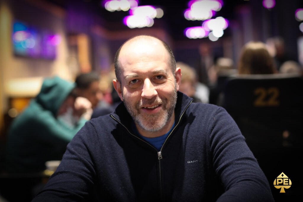 Ein Mann in einem schwarzen Anzug und einer roten Krawatte spielt konzentriert Poker an einem grünen Pokertisch mit Chips und Karten vor sich.