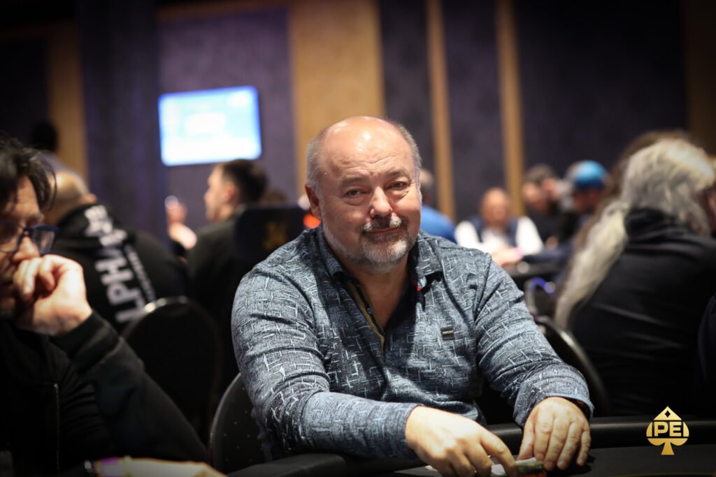 Ein Mann und eine Frau sitzen an einem Pokertisch in einem luxuriösen Casino. Der Mann hat Karten in der Hand und beide haben konzentrierte Gesichtsausdrücke. Im Hintergrund sind andere Spieler