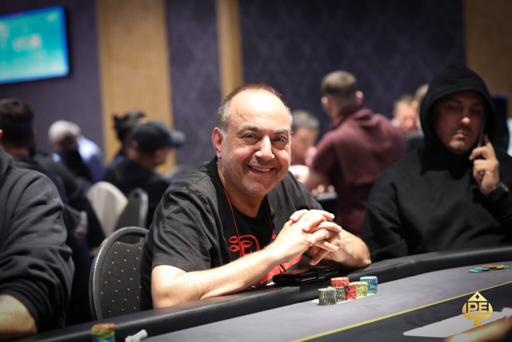 Ein Mann in einem schwarzen Anzug und einer weißen Hemd steht vor einem Pokertisch mit Chips und Karten. Er hält zwei Spielkarten in der Hand und zeigt ein selbstbewusstes Lächeln.