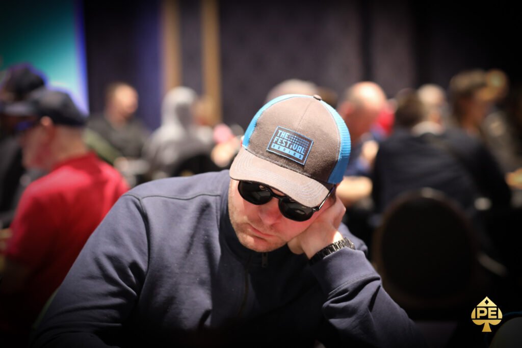 Ein Mann sitzt an einem Pokertisch und hält zwei Karten in der Hand, während er konzentriert auf seine Gegner schaut. Eine Reihe von Pokerchips liegt vor ihm auf dem Tisch.
