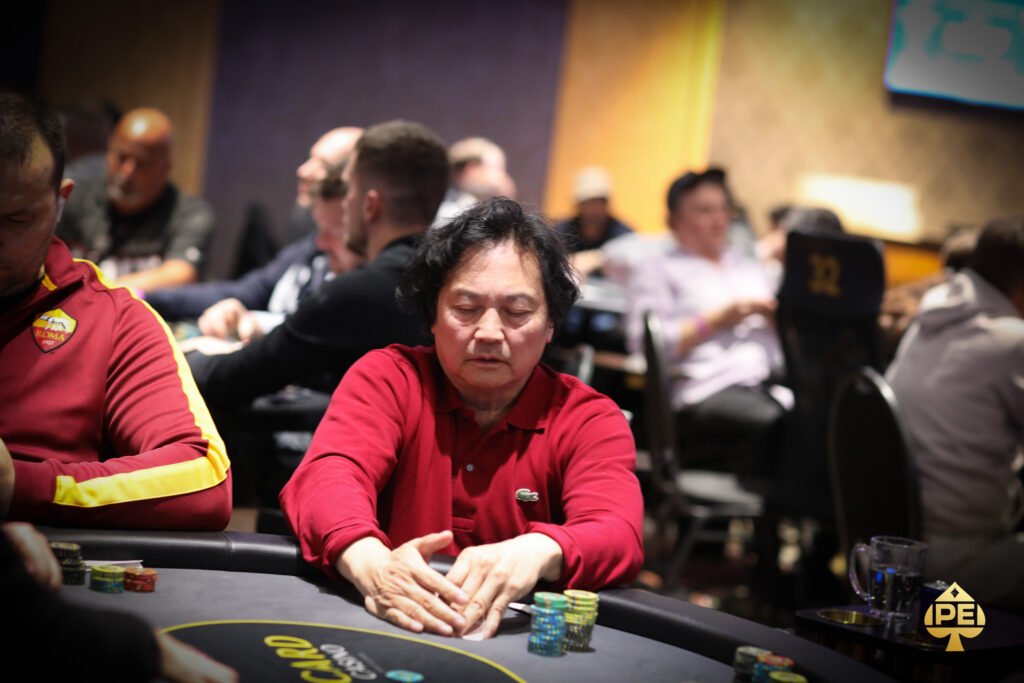 Ein Mann sitzt an einem Pokertisch und hält Karten in der Hand, während er konzentriert auf seine Gegner schaut. Die Szene spielt in einem eleganten Casino mit stilvoller Beleuchtung und