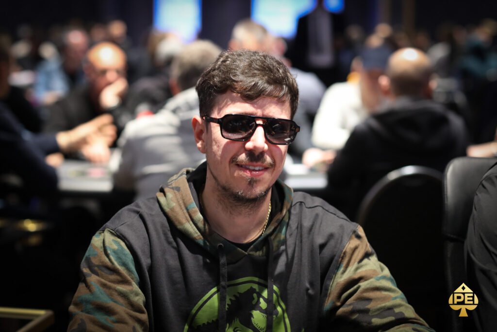 Ein Mann in einem dunkelblauen Anzug und weißem Hemd sitzt an einem Pokertisch und hält seine Karten in der Hand. Im Hintergrund sind weitere Spieler zu sehen, die gespannt auf die Karten