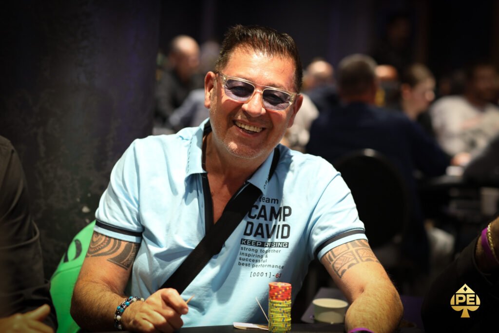 Ein Mann sitzt an einem Pokertisch und hält zwei Spielkarten in der Hand. Er hat einen konzentrierten Blick und trägt ein dunkles Hemd. Im Hintergrund sind weitere Spieler und Pokerchips zu sehen