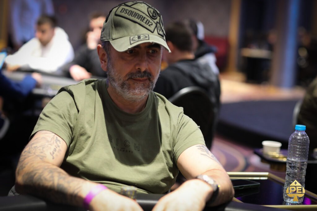 Ein Mann in einem dunkelblauen Anzug sitzt an einem Pokertisch und hält eine Hand mit Spielkarten. Er zeigt ein selbstbewusstes Lächeln, während er seinen Gegnern gegenüberblickt.
