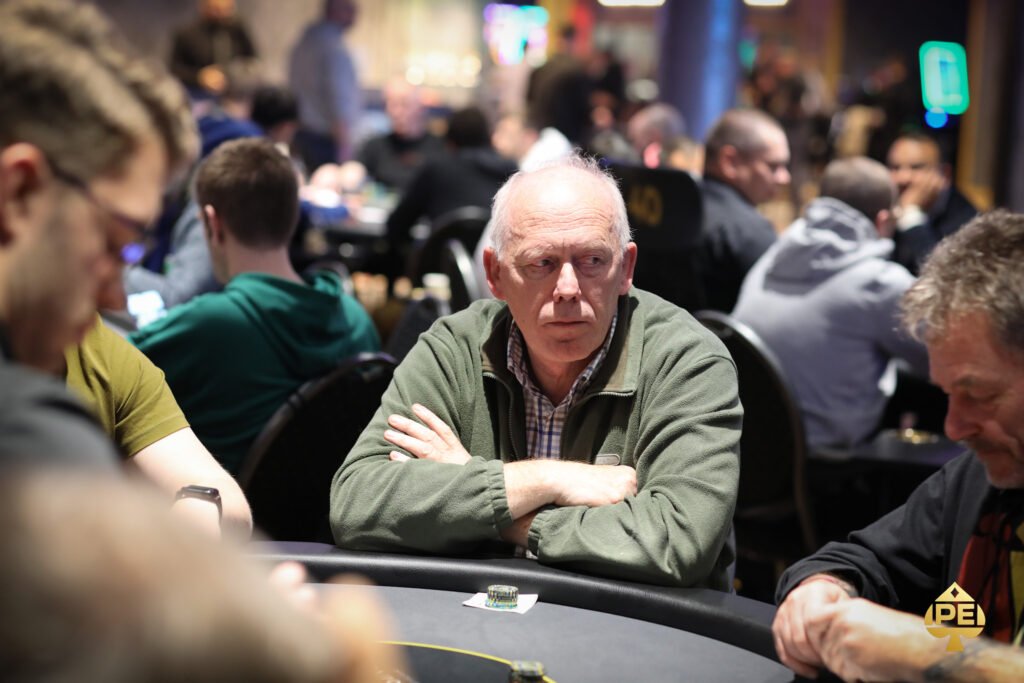 Ein Mann in einem dunkelblauen Anzug und einer weißen Krawatte sitzt an einem Pokertisch und hält seine Spielkarten in der Hand. Im Hintergrund sind andere Spieler und Chips auf dem Tisch zu sehen