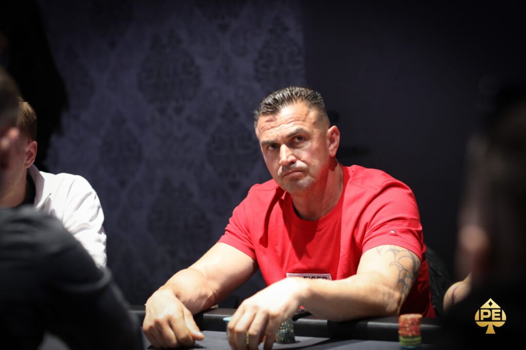 Ein Mann sitzt an einem Pokertisch mit grüner Filzdecke und hält zwei Spielkarten in der Hand. Er hat konzentrierten Blick auf die Karten und trägt eine schwarze Kappe sowie ein dunk
