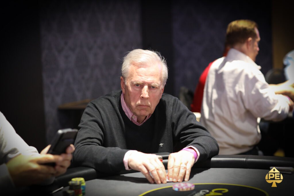 Ein junger Mann spielt Poker an einem eleganten Tisch in einem luxuriösen Casino. Er hält Karten in der Hand und betrachtet konzentriert die Gemeinschaftskarten auf dem Tisch. Andere
