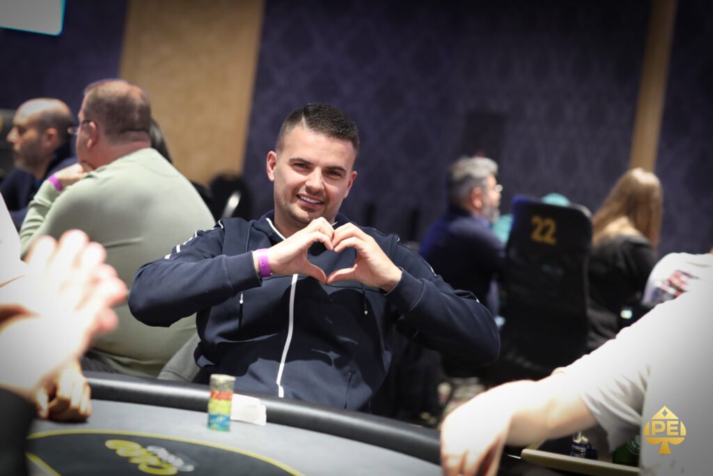Ein Mann in einem dunkelblauen Anzug und einer roten Krawatte sitzt an einem Pokertisch und hält eine Hand mit Spielkarten. In der Mitte des Tisches liegen weitere Karten. Hinter ihm