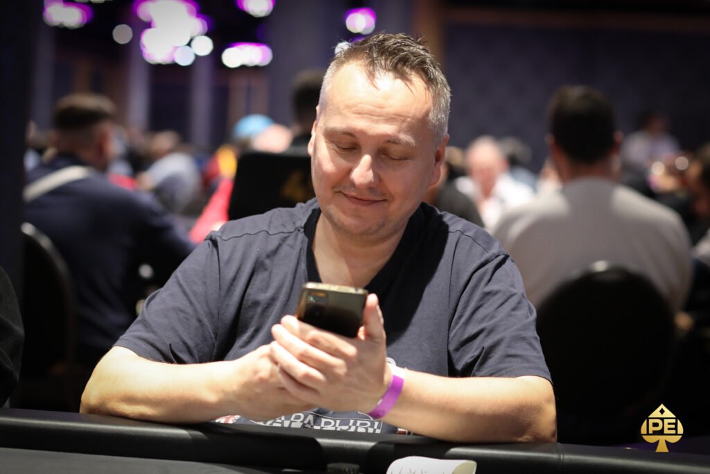 Ein Mann sitzt an einem Pokertisch, hält Spielkarten in der Hand und schaut konzentriert auf den Tisch. Im Hintergrund sind andere Spieler und Casino-Chips zu sehen.