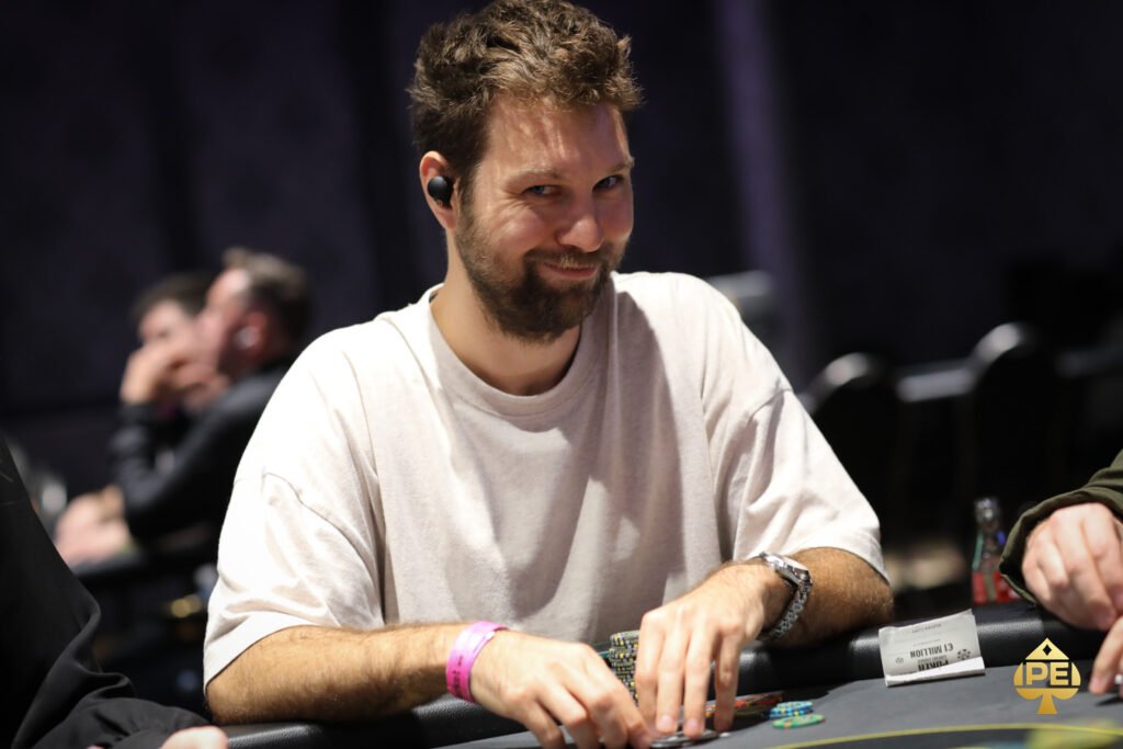 Ein Mann in einem Anzug und mit Sonnenbrille sitzt an einem Pokertisch und hält eine Hand mit Spielkarten. Andere Spieler sind im Hintergrund zu sehen.