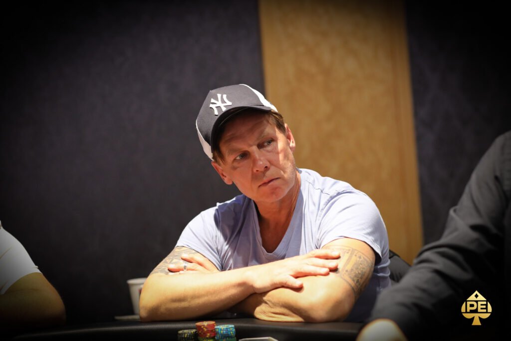 Ein Mann sitzt an einem Pokertisch und hält zwei Karten in der Hand. Er trägt einen Anzug und hat eine ernste Miene. Im Hintergrund sind weitere Spieler und Chips zu sehen.