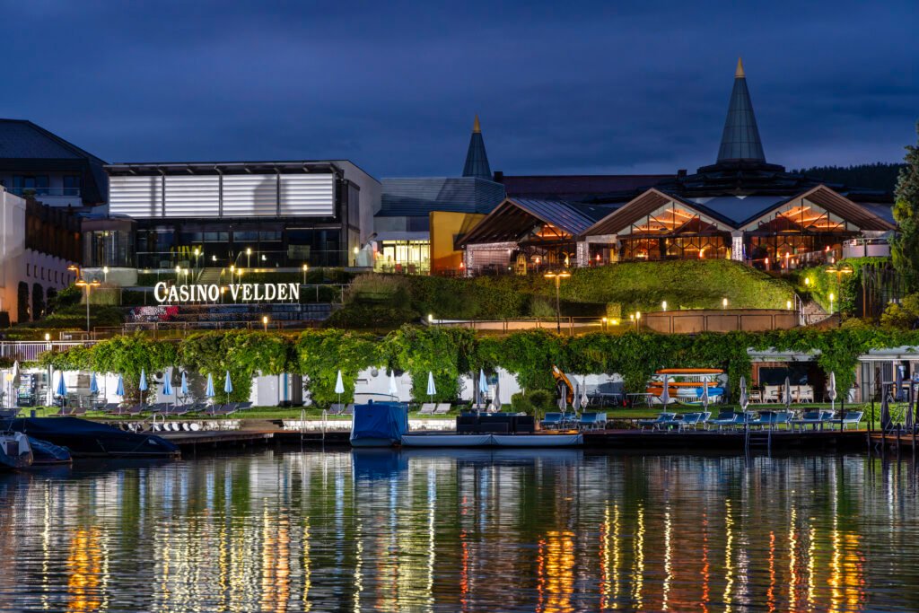Ein nächtliches Bild von Velden am Wörthersee. Die beleuchtete Promenade spiegelt sich im ruhigen Wasser des Sees wider, während im Hintergrund die Lichter der Stadt funkeln.