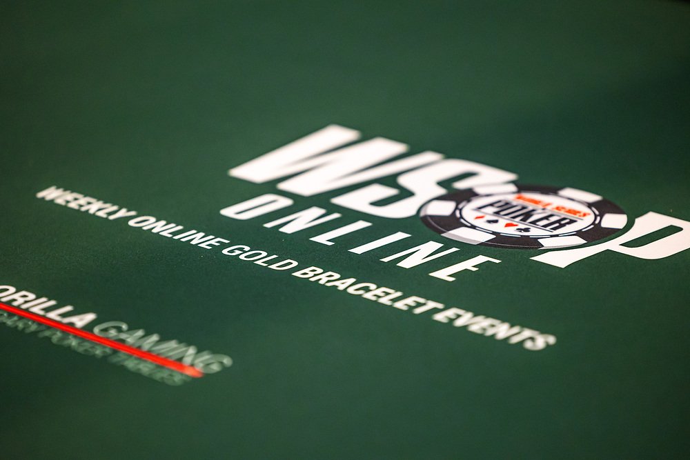 Ein professioneller Pokerspieler mit Sonnenbrille und Kappe sitzt an einem Tisch während des World Series of Poker-Turniers. Er hält Karten in der Hand und betrachtet konzentriert seine Gegner